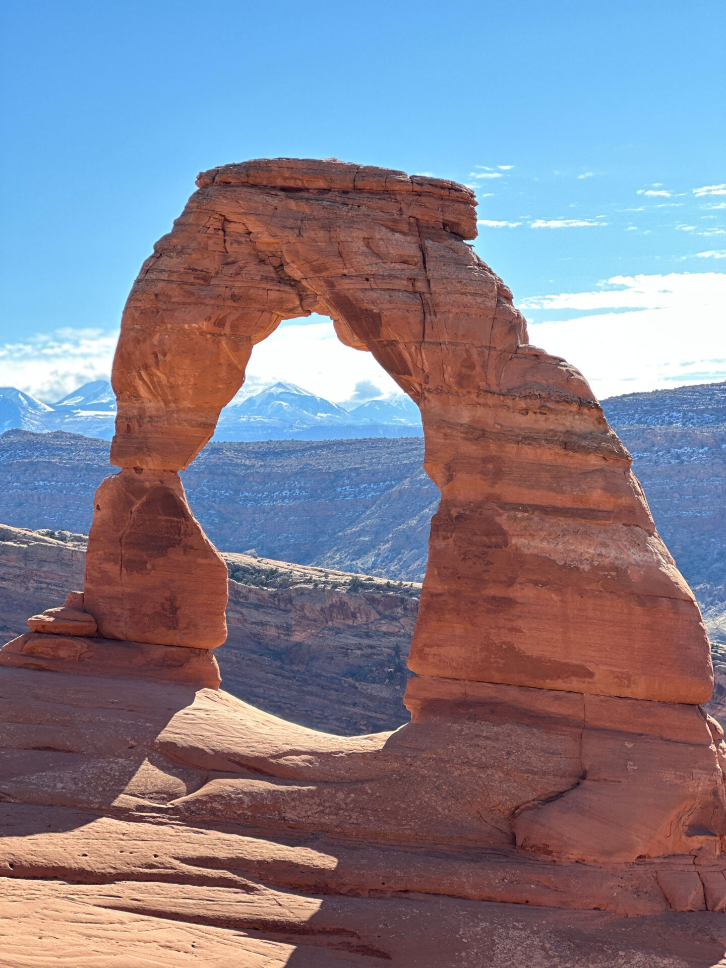 Delicate Arch, a towering sandstone arch in Arches National Park, Utah, soars gracefully above a sloping desert landscape. The arch is approximately 46 feet tall and 32 feet wide, with a delicate appearance that defies its massive size. Its smooth, rounded surface is a testament to the millions of years of erosion that have shaped it. In the background, a range of rugged mountains rises up, their peaks capped with snow. The sky is a clear blue, with a few white clouds floating by.